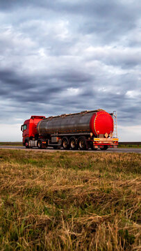 Red semi truck with a silver tanker trailer moving on an asphalt highway, against background of cloudy sky and foreground of dry grass, representing liquid cargo transportation vertical photo