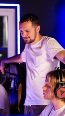 Obraz premium Young man with beard, casual white t-shirt, standing in computer club with glowing blue neon light background, observing activity of seated person, indoors environment vertical photo
