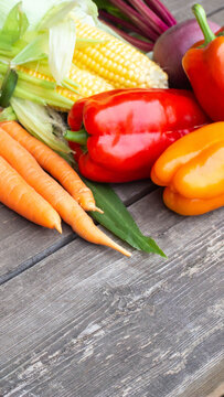 Assortment of vibrant, colorful fresh harvest vegetables including yellow corn on cob, red and orange bell peppers, and orange carrots arranged on rustic wooden table, overhead view vertical photo