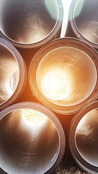 Close-up view of large corrugated plastic pipes stacked outdoors, bright sunlight illuminating pipe interiors, lens flare effect, construction materials, infrastructure development vertical photo