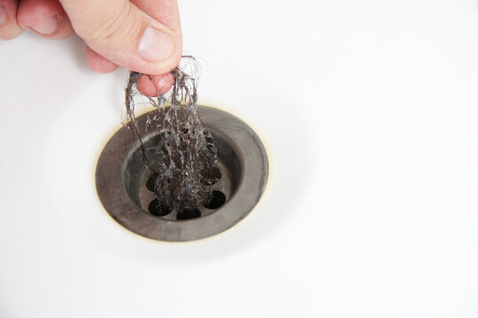 Person hand pulling tangled hair mass from white bathroom sink drain, illustrating common plumbing issue, hygiene maintenance, and household problem solution for clogged pipes, copy space