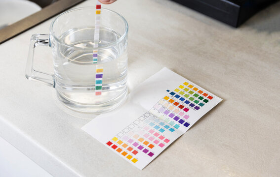 Hand of person immersing water quality test strip in glass of clear water to measure parameters on stone surface, analysis of chemical composition of liquid with special tool and color chart