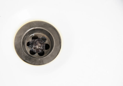 Close-up view of dark hair mass caught in metallic drain of white bathroom sink causing plumbing obstruction problem home maintenance issue needing repair, copy space