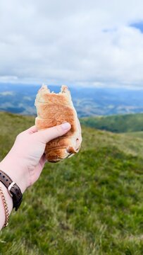 Person Holding Bitten Croissant Sandwich with Scenic Mountain Landscape Background