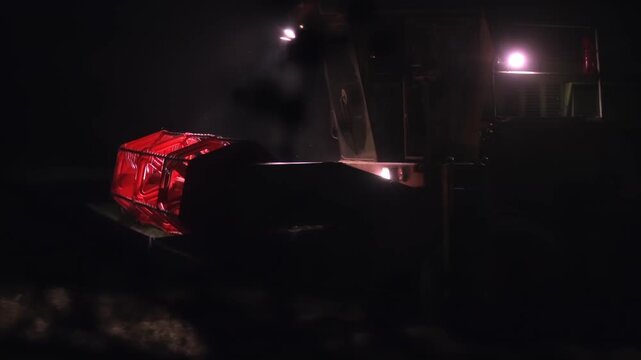 A combine harvester cuts barley at night, moving through a dark field with its red header and bright work lights. Illuminated stubble and rising dust create a dramatic, cinematic remote harvest scene.