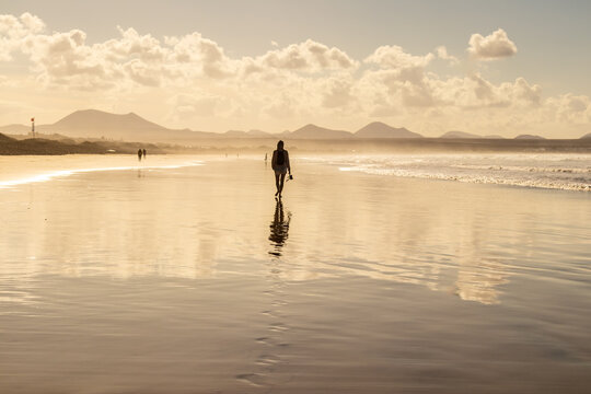 Woman walking barefoot along the wet sandy shore of Famara beach in Lanzarote, Canary Islands, holding her sandals. Scenic coastal landscape with ocean reflections and vacation vibe.