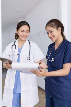 Two young Asian female medical professionals, a doctor in a white coat and a nurse in scrubs and reviewing data on a tablet and clipboard in a hospital hallway.