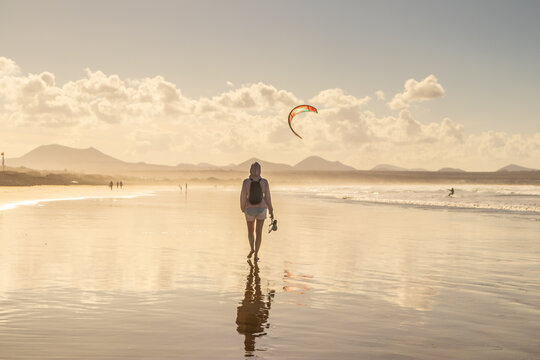 A woman walking barefoot along the wet sandy shore of Famara beach in Lanzarote, Canary Islands, holding her sandals. Scenic coastal landscape with ocean reflections and vacation vibe.