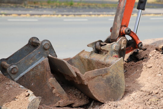 Excavator bucket digging a trench beside a temporary safety barrier during road construction. Heavy equipment working on street infrastructure maintenance.