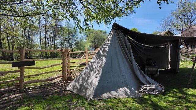 Historical canvas tent pitched on green grass in a forest settlement, illuminated by bright sunlight with dappled shadows, featuring a wooden log fence, an open shelter with clay pots, and thatched hu