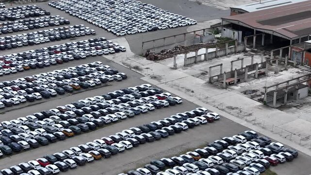 Large automotive logistics yard filled with thousands of parked passenger vehicles at a vehicle distribution terminal, showing organized storage rows and industrial facilities drone lateral slide