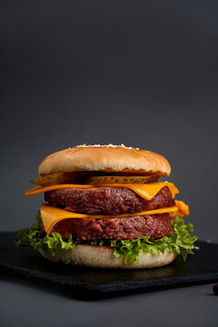 Juicy double cheeseburger with two meat patties, melted cheddar, pickles, and lettuce on black slate board, dark background.