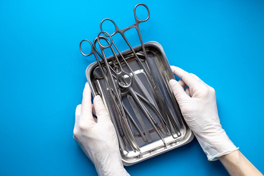 Hand in white glove is holding sterile surgical instruments and tools - scalpels, scissors and others - in a stainless tray, top view