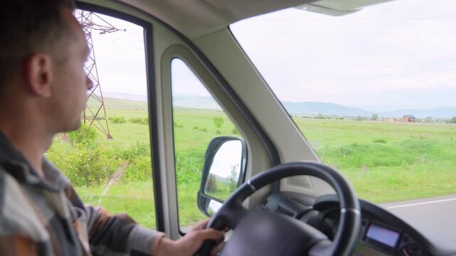 Serious man driving a camper van on a rural asphalt road, observing the beautiful green landscape from the window while holding the steering wheel and enjoying his summer vacation travel