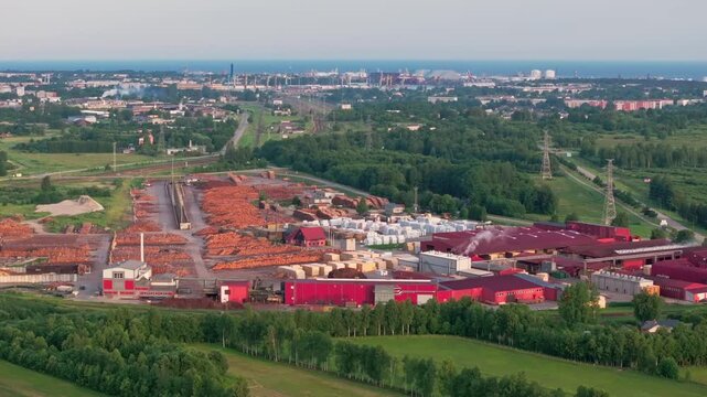 Aerial drone footage of a large timber processing and wood manufacturing facility near Ventspils, Latvia, with the Port of Ventspils visible in the background at sunrise. Expansive log storage yards,