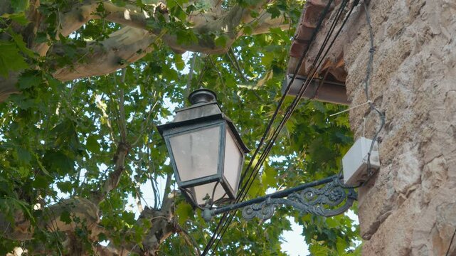 An ornate wrought iron lantern mounted on a rustic stone wall beneath green plane tree branches. Classic Mediterranean village detail in soft light. Medium shot.