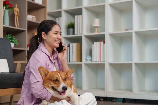 Asian woman talking on smartphone while holding her corgi dog at home