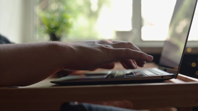 Close-up of hands typing on a laptop keyboard with a blurred window background