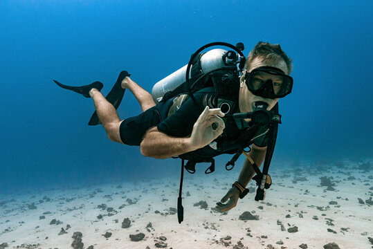 Caucasian male Scuba diver doing the hand signal for all is ok underwater, with sandy bottom and blue water. 