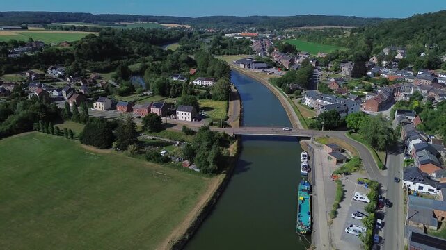 Aerial view of Namur's Sambre River flowing through green fields and town. Bridge crosses calm water near parked boats and houses. Lush forests and farmland surround peaceful village
