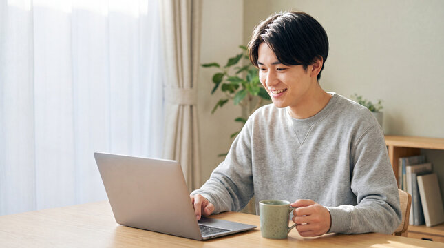 明るい室内でパソコン作業をしながら微笑む若い男性 ｜ 自然光の入る部屋
