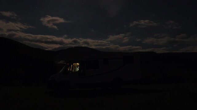 A bright full moon moving across the night sky, illuminating the clouds over a motorhome parked in a tranquil mountain valley, symbolizing freedom, adventure, and nomadic life