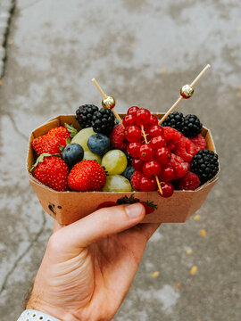 Close-up of a hand holding a cardboard tray filled with fresh berries and grapes purchased at the Central Market in Ljubljana, Slovenia.