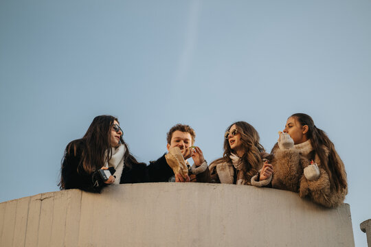 Four casual friends stand on a rooftop ledge wearing winter coats while sharing snacks and laughing. A group of young people enjoy a fun outdoor moment together against a clear sky.
