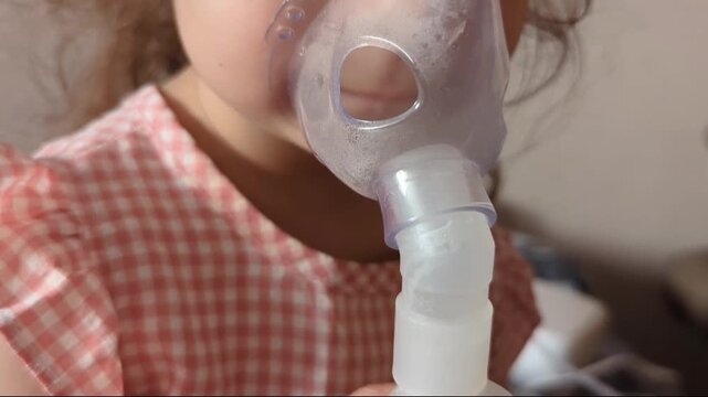 young child wearing a clear medical nebulizer mask over their nose and mouth for inhalation therapy, clad in a pink checkered dress in an indoor setting.
