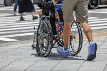 Fototapeta premium Rear view of a man pushing a wheelchair user across a city crosswalk with pedestrians and city traffic in the background.