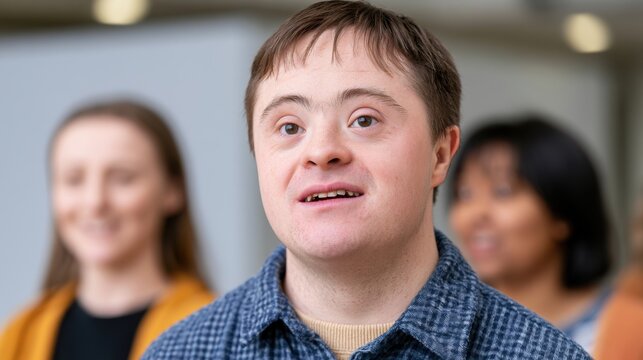 Young male with Down syndrome wearing a blue checkered shirt smiles while standing in a group of diverse individuals in a bright indoor setting
