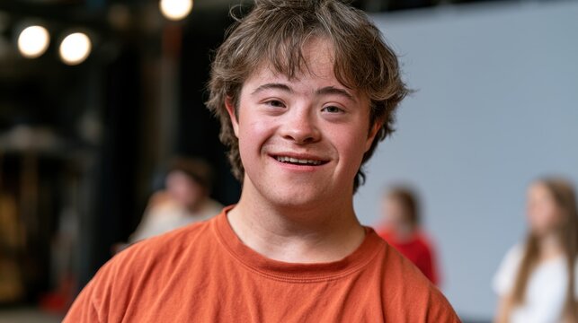 Young male with Down syndrome wearing an orange t-shirt smiles at the camera in a brightly lit indoor setting with blurred figures in the background