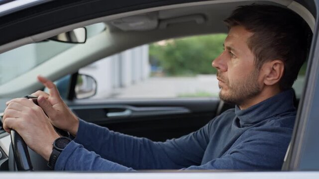 Man waiting behind the steering wheel, looking around, taps on the wheel and checks his wristwatch while sitting inside a parked car