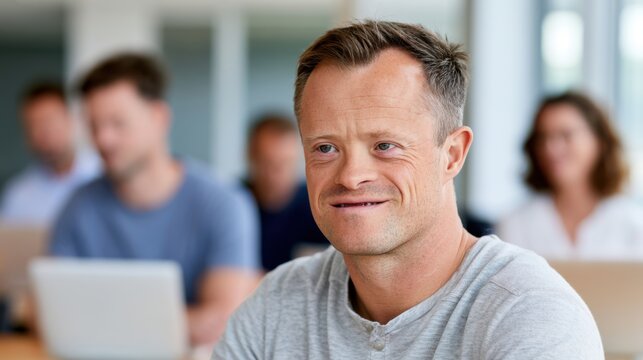 Adult male with Down syndrome smiles while seated at a table in a modern office environment, with blurred colleagues and laptops in the background