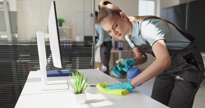 Cleaner woman cleaning office desk with spray. Professional janitor team ensures service and hygiene in an office, sanitizing equipment and wiping surfaces. Office cleaning and hygiene theme.
