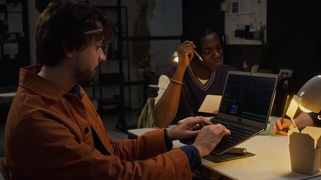 Multiethnic young adult programmers working late in office, Caucasian man typing code on laptop while African American colleague speaking. Suitable for software development and IT startup ads