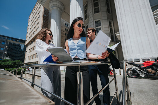Three business professionals stand by a railing studying reports and charts. The team reviews documents together outside a classical office building for a fast outdoor meeting.