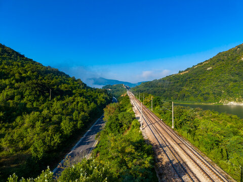 Train tracks run parallel to the river surrounded by green hills on a clear day with blue skies
