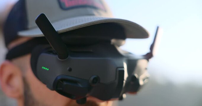 FPV drone operator wears digital goggles under a cap in harsh sunlight, green LEDs glowing on the headset.