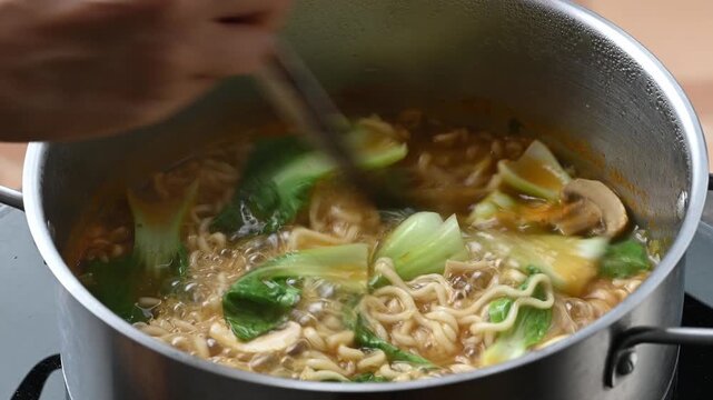 Stirring spicy instant noodle soup with bok choy, mushrooms, quail eggs and tteokbokki in a boiling pot using chopsticks