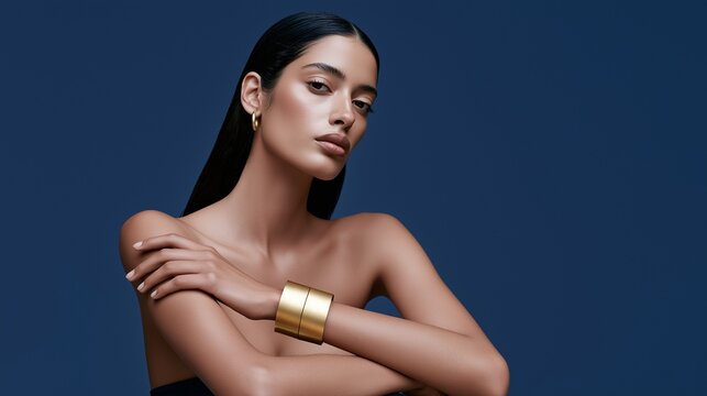 Young woman with sleek black hair crosses her arms over bare shoulders while wearing a wide gold cuff, suited to jewelry, skincare, self-care and luxury fashion campaigns.