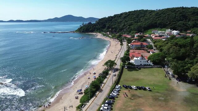 Aerial drone footage following the coastline of Praia da Saudade in Penha, Santa Catarina, Brazil. The view shows a sandy beach, blue-green Atlantic waters, rocky formations.