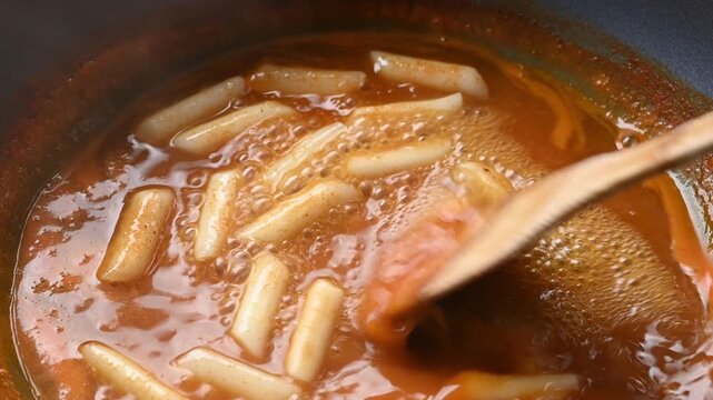 Stirring Korean Tteokbokki (rice cake) in spicy red sauce in a hot pan with a wooden spatula