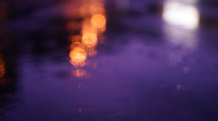Fototapeta premium A blurred abstract image featuring warm orange bokeh lights reflecting on a dark purple wet surface, creating a dreamy and atmospheric effect.