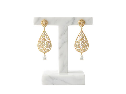 A pair of ornate gold teardrop earrings with filigree patterns and a single clear stone drop displayed on a white marble t-shaped jewelry stand isolated on a transparent background