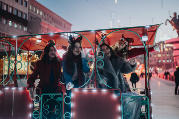 Fototapeta premium Four young women laugh together on a brightly lit festive ride at a Christmas market. The friends celebrate the holiday season on a winter evening with lights and decorations.