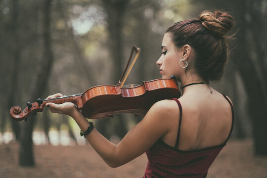 Rear view of a cool teenage girl standing in a forest playing the violin