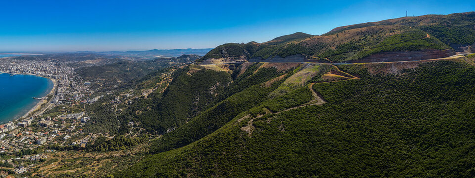 Breathtaking drone footage capturing a winding coastal highway carved into a steep, lush green mountainside, overlooking the expansive blue bay and distant cityscape of Vlora, Albania.