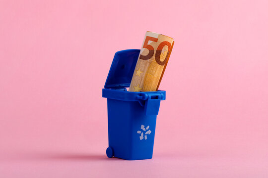 Fifty euro banknote in blue trash bin on pink background, symbolizing financial waste or poor spending decisions.