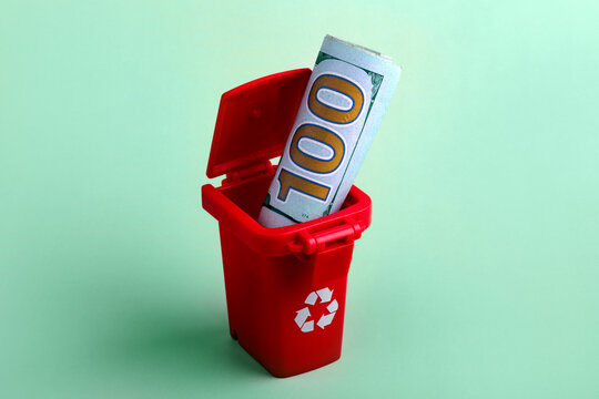 Rolled hundred dollar bill placed inside a small red recycling bin on a mint green background, symbolizing wasteful spending or financial loss.